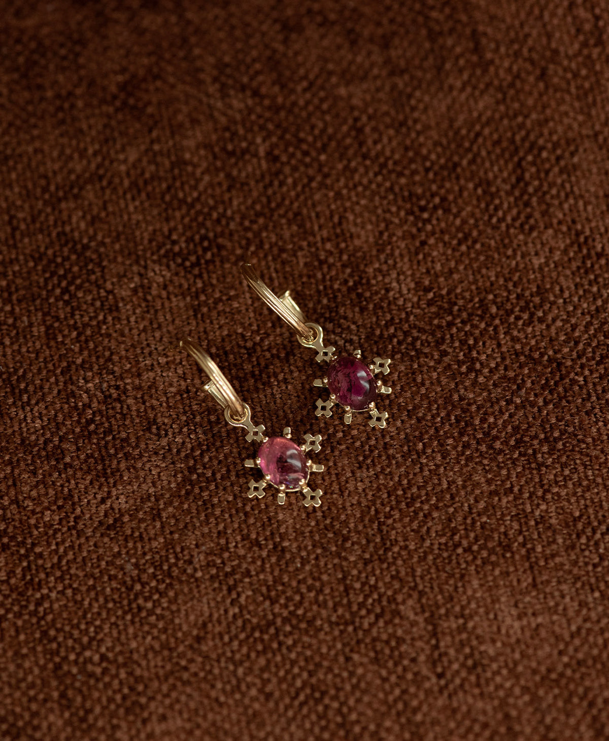 A pair of Farah gold drop earrings with pink tourmaline gemstones, displayed on a textured brown fabric background.