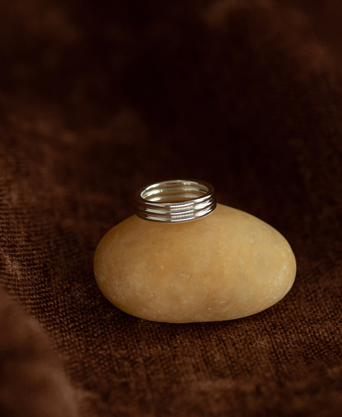 The silver Irma Stacker Ring, formed from three connected bands, layered closely, rests on a cream stone on a textured brown background.