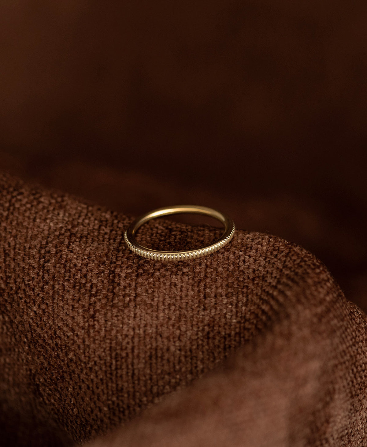 The gold Kenza Stacking Ring with fine engraved lines encircling the band, rests on a textured brown background.