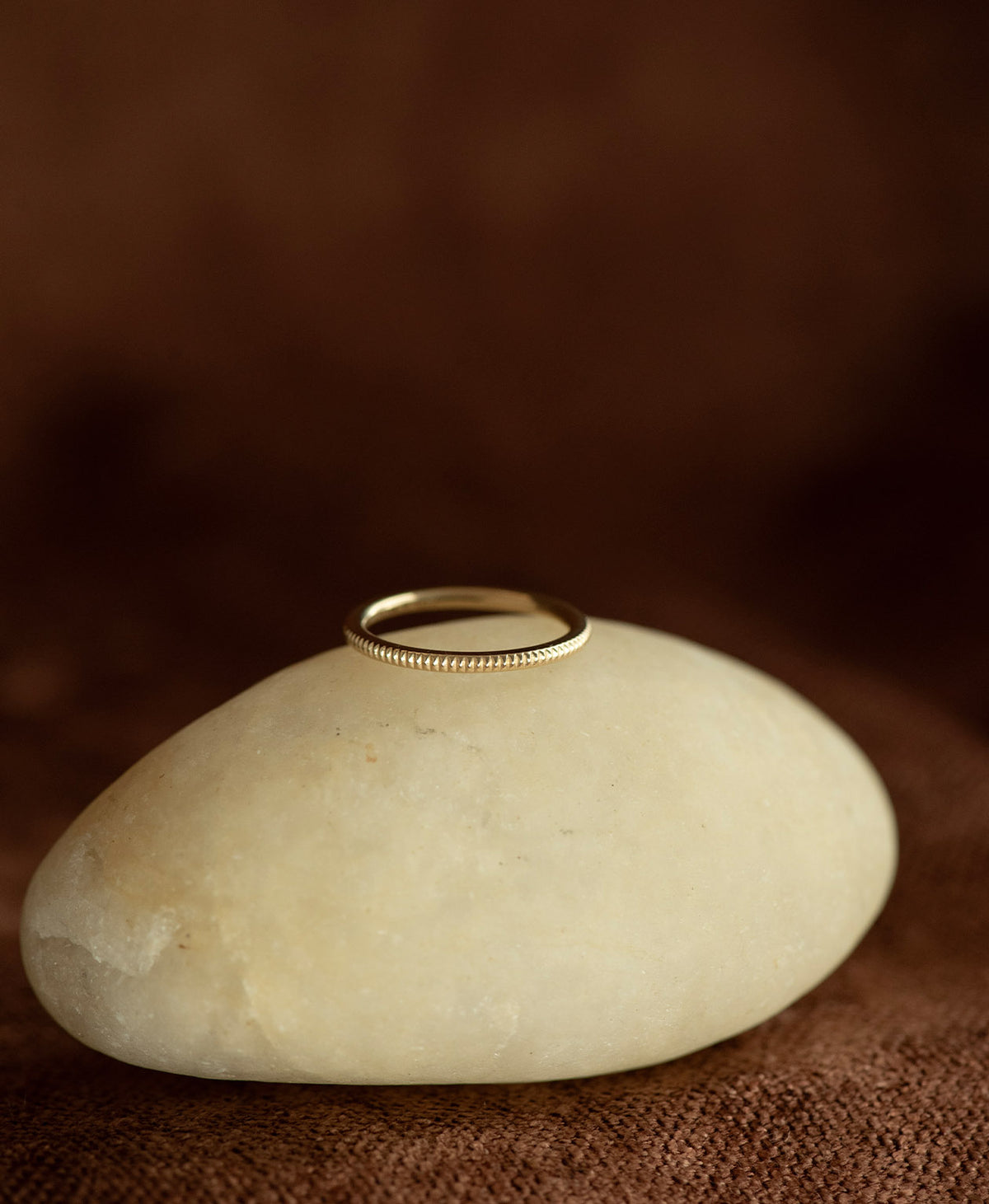 The gold Kenza Stacking Ring with fine engraved lines encircling the band, rests on a cream stone on a textured brown background.