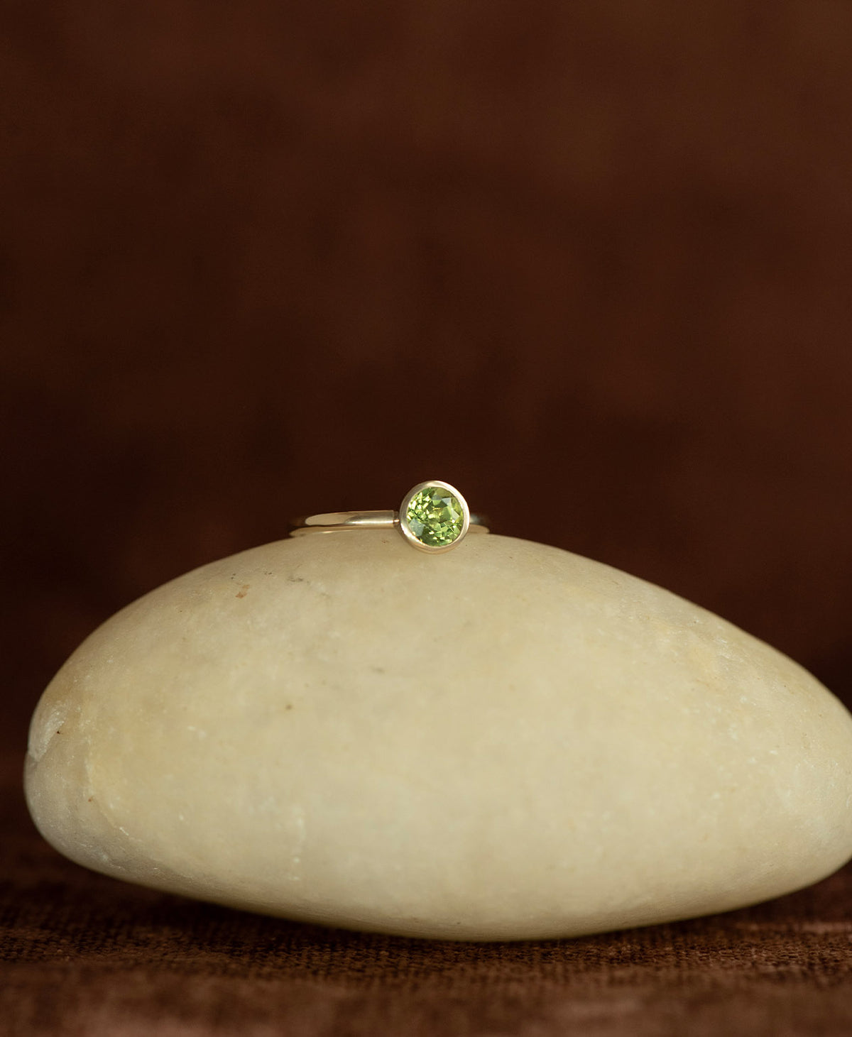 The gold Mon Petit Stacker Ring with a bezel set, round cut Peridot rests on a cream stone on a textured brown background.