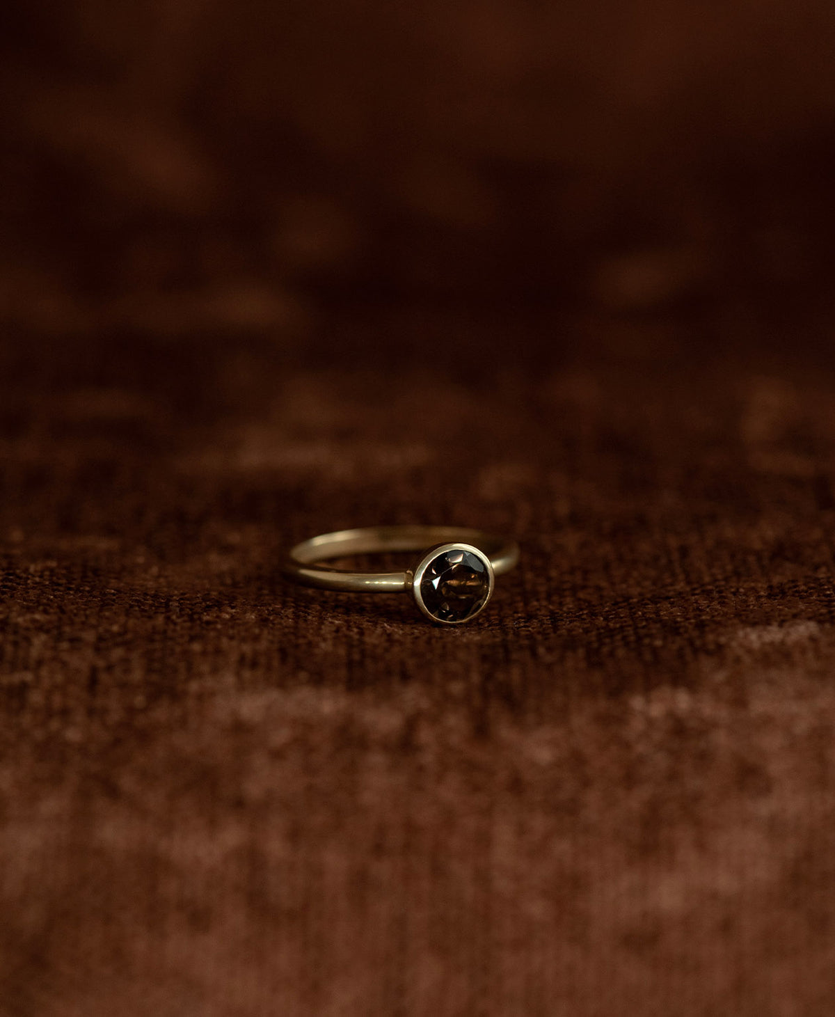 The gold Mon Petit Stacker Ring with a bezel set, round cut smoky quartz rests on a textured brown background. 