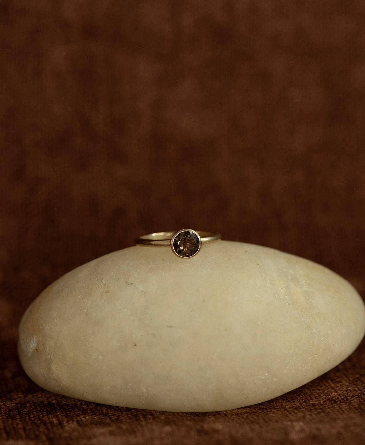 The gold Mon Petit Stacker Ring with a bezel set, round cut smoky quartz rests on a cream stone on a textured brown background. 