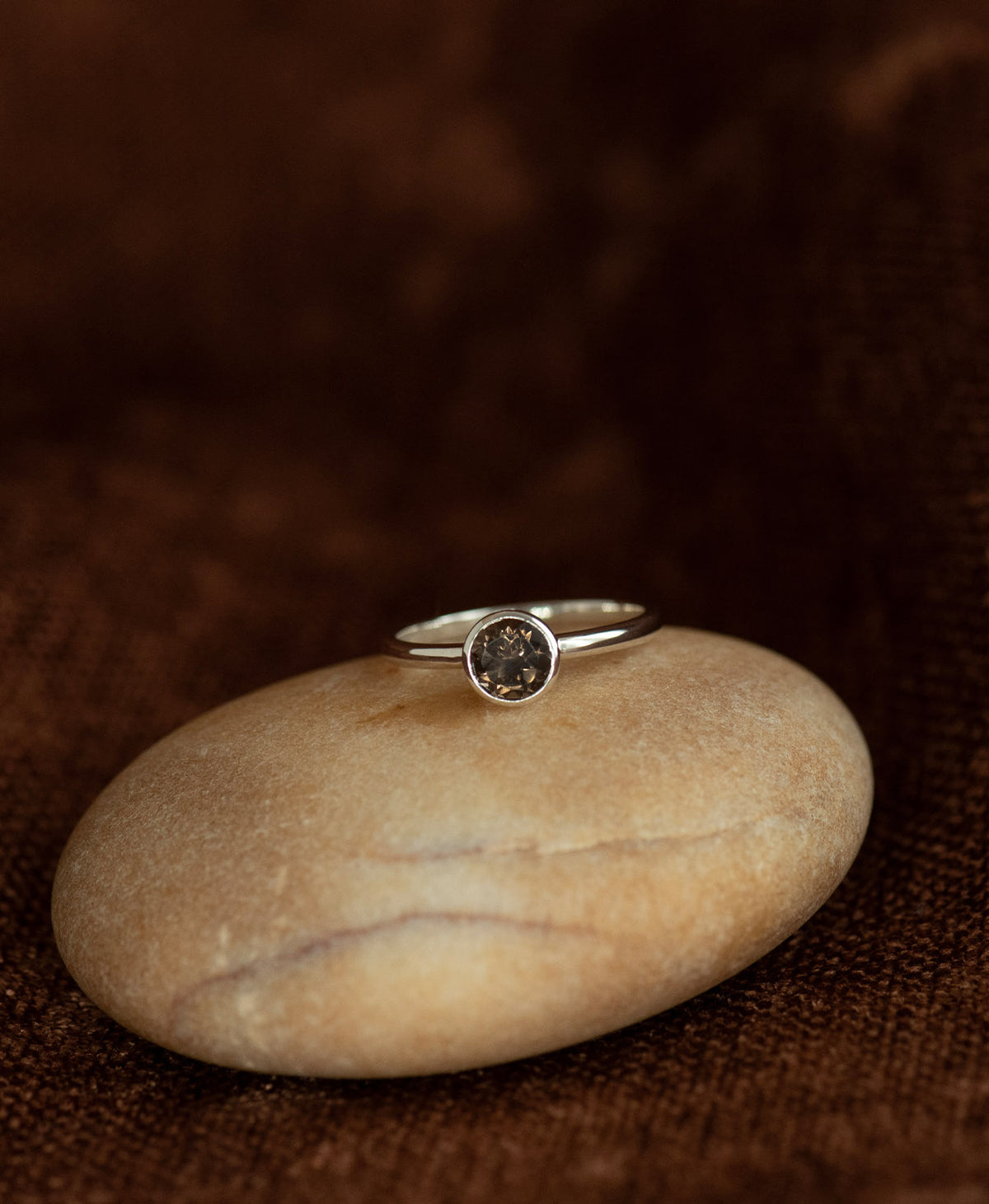 A close up of the silver Mon Petit Stacker Ring with a bezel set, round cut smoky quartz resting on a cream stone on a textured brown background. 