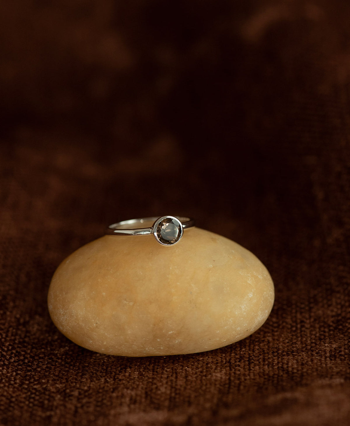 The silver Mon Petit Stacker Ring with a bezel set, round cut smoky quartz rests on a cream stone on a textured brown background. 