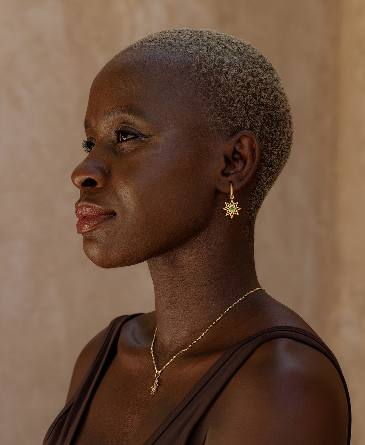 A close-up profile of a person wearing the Nora Hoops, featuring a gold star hanging from a hoop with a central green stone, paired with a gold necklace with a star pendant.