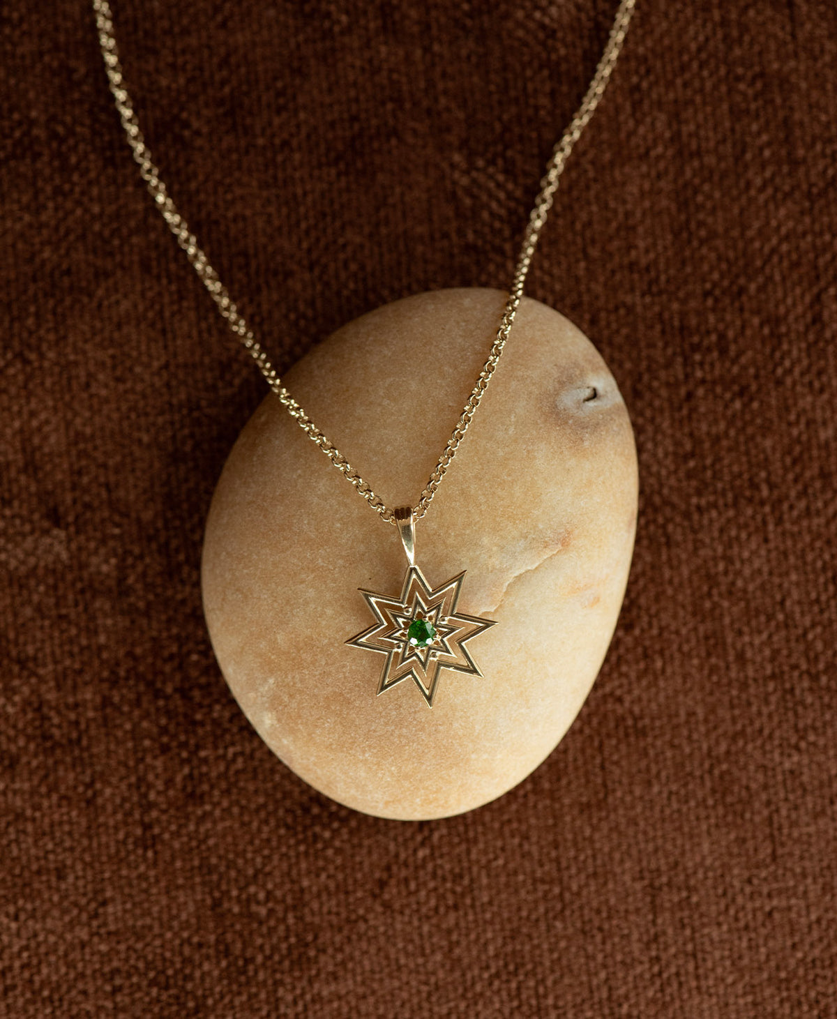 The Nora Necklace featuring a gold starburst pendant necklace with a small chrome diopside at its centre, displayed over a cream stone on a textured brown fabric background.
