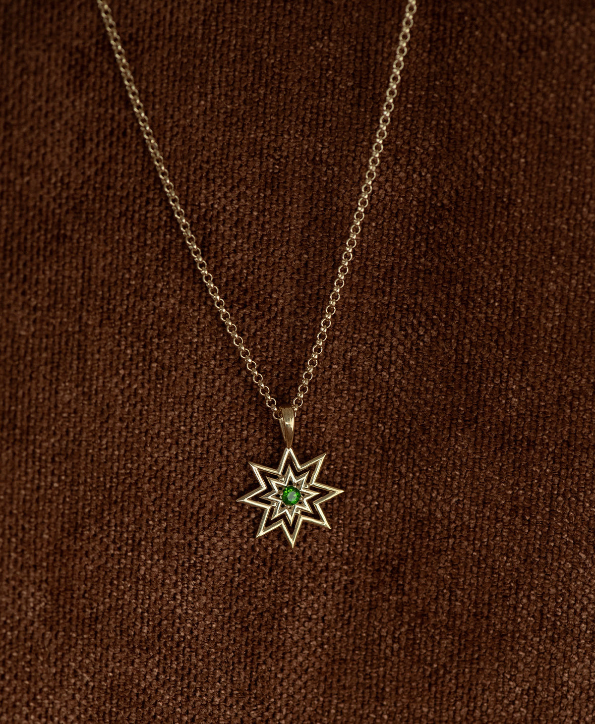 The Nora Necklace featuring a gold starburst pendant necklace with a small chrome diopside at its centre, displayed on a textured brown fabric background.