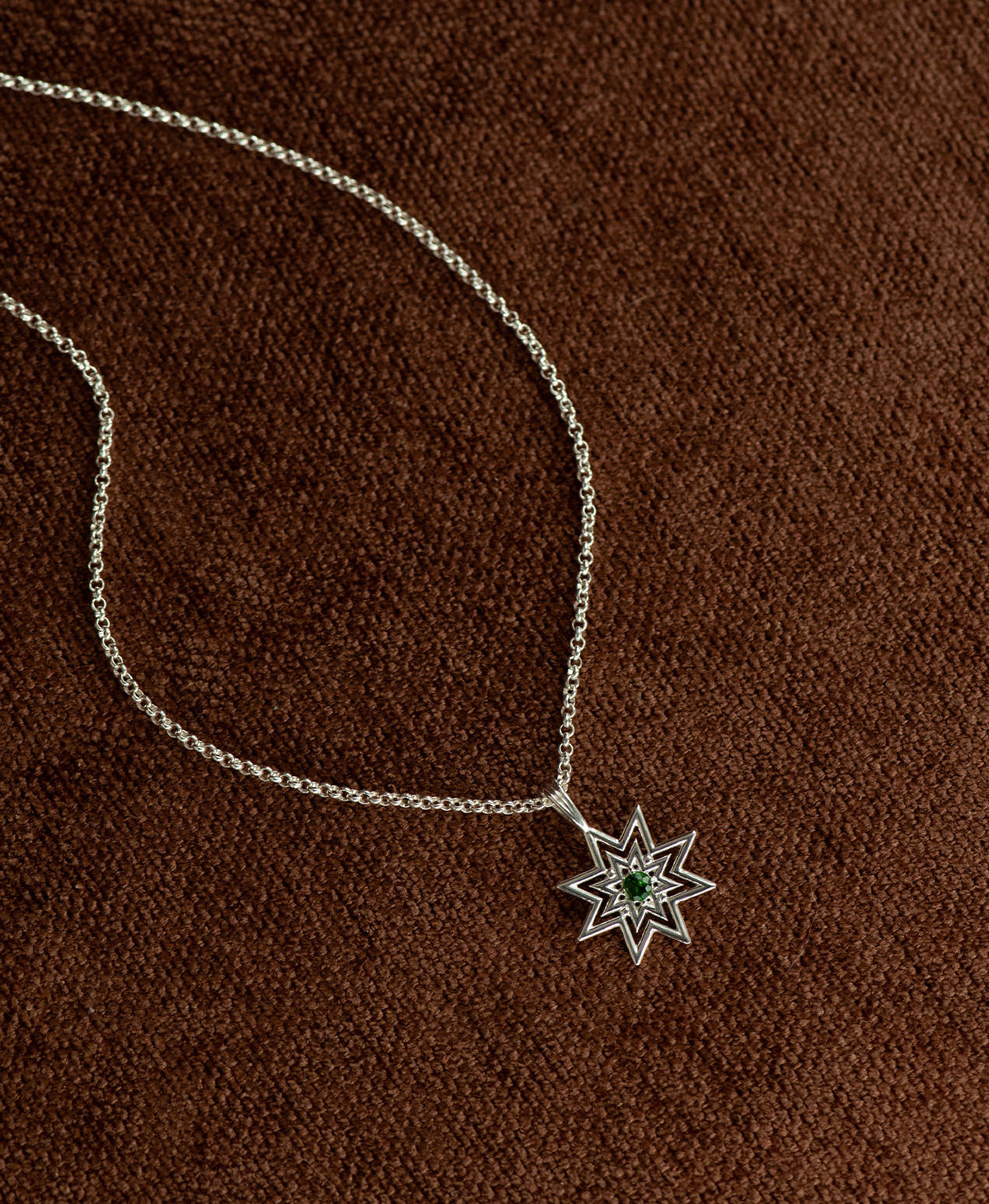 The Nora Necklace featuring a silver starburst pendant necklace with a small chrome diopside at its centre, displayed on a textured brown fabric background.