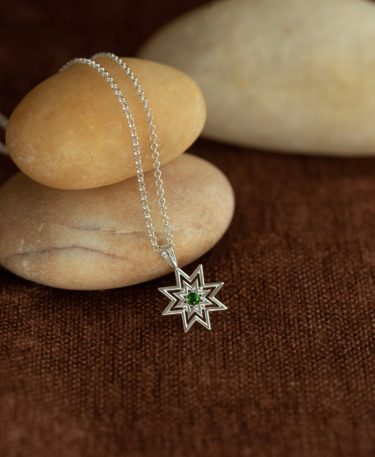 The Nora Necklace featuring a silver starburst pendant necklace with a small chrome diopside at its centre, displayed over a stack of cream stones on a textured brown fabric background.