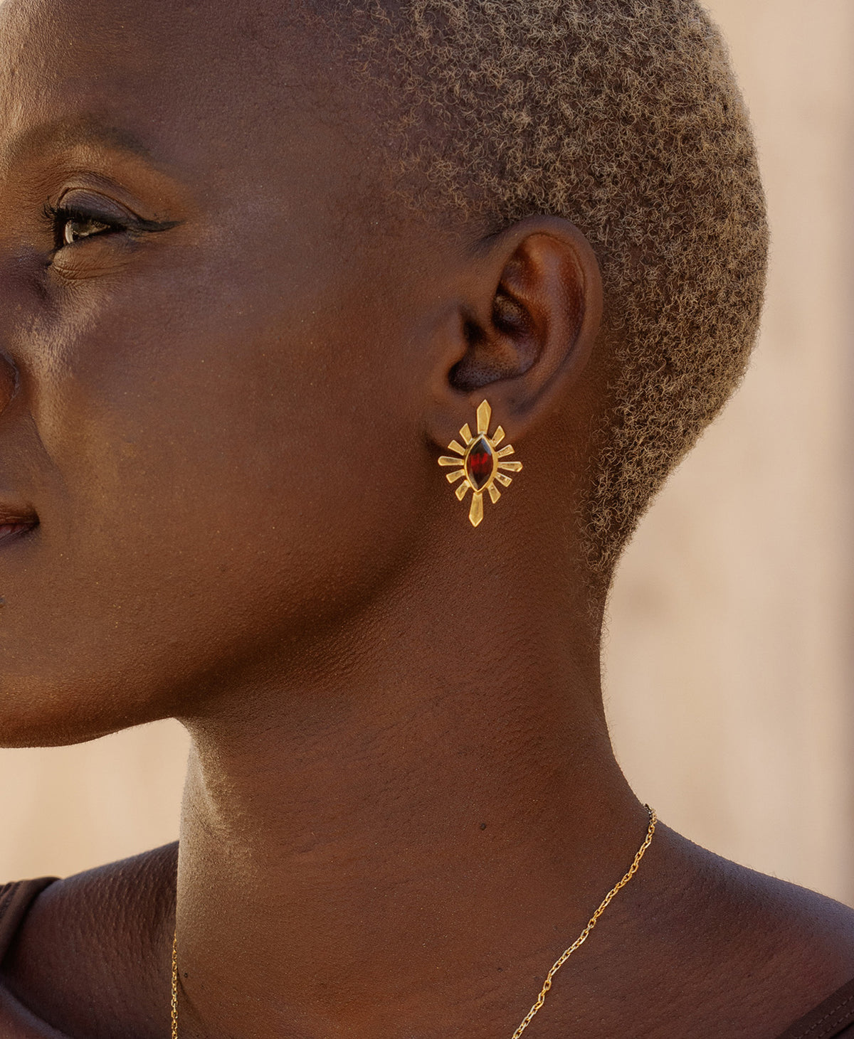 A close-up side profile of a person wearing the Sister Studs, featuring a marquise cut garnet in a gold linear sunburst frame. 