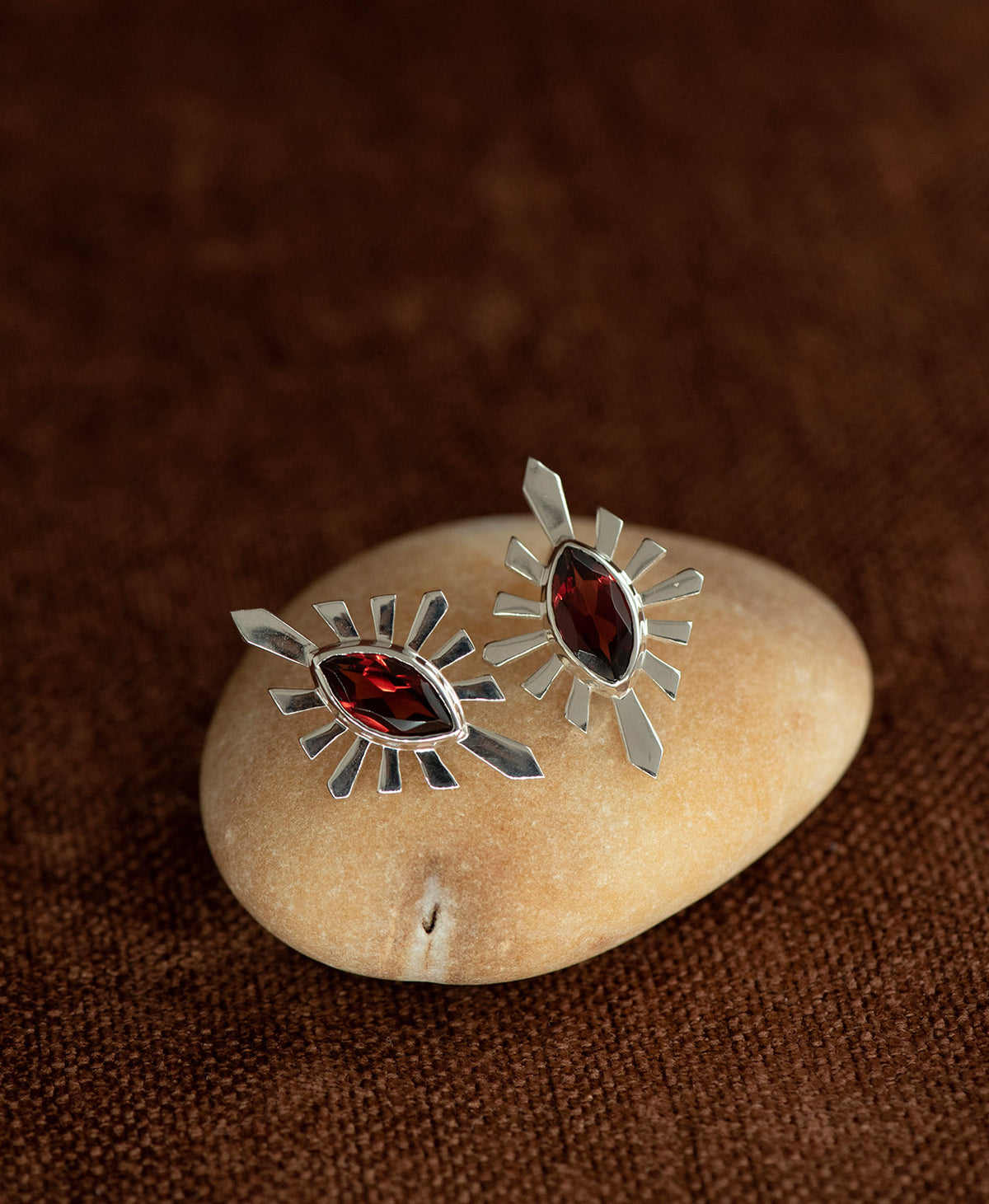 The Sister Studs, featuring a central marquise cut Garnet nestled within a silver sunburst frame, displayed on a smooth beige stone against a textured brown fabric background.