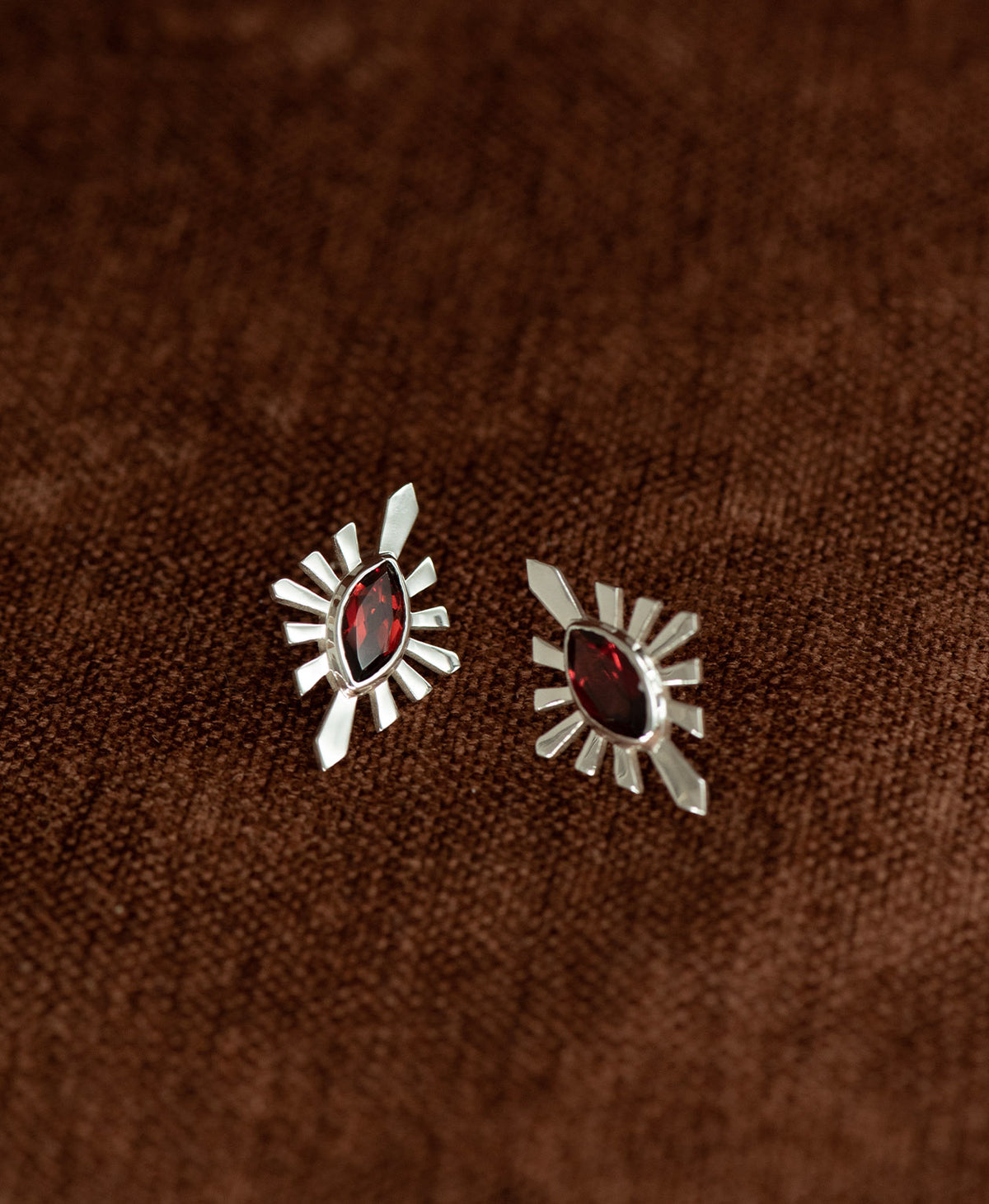 The Sister Studs, featuring  a central marquise cut Garnet nestled within a silver sunburst frame, displayed against a textured brown fabric background.