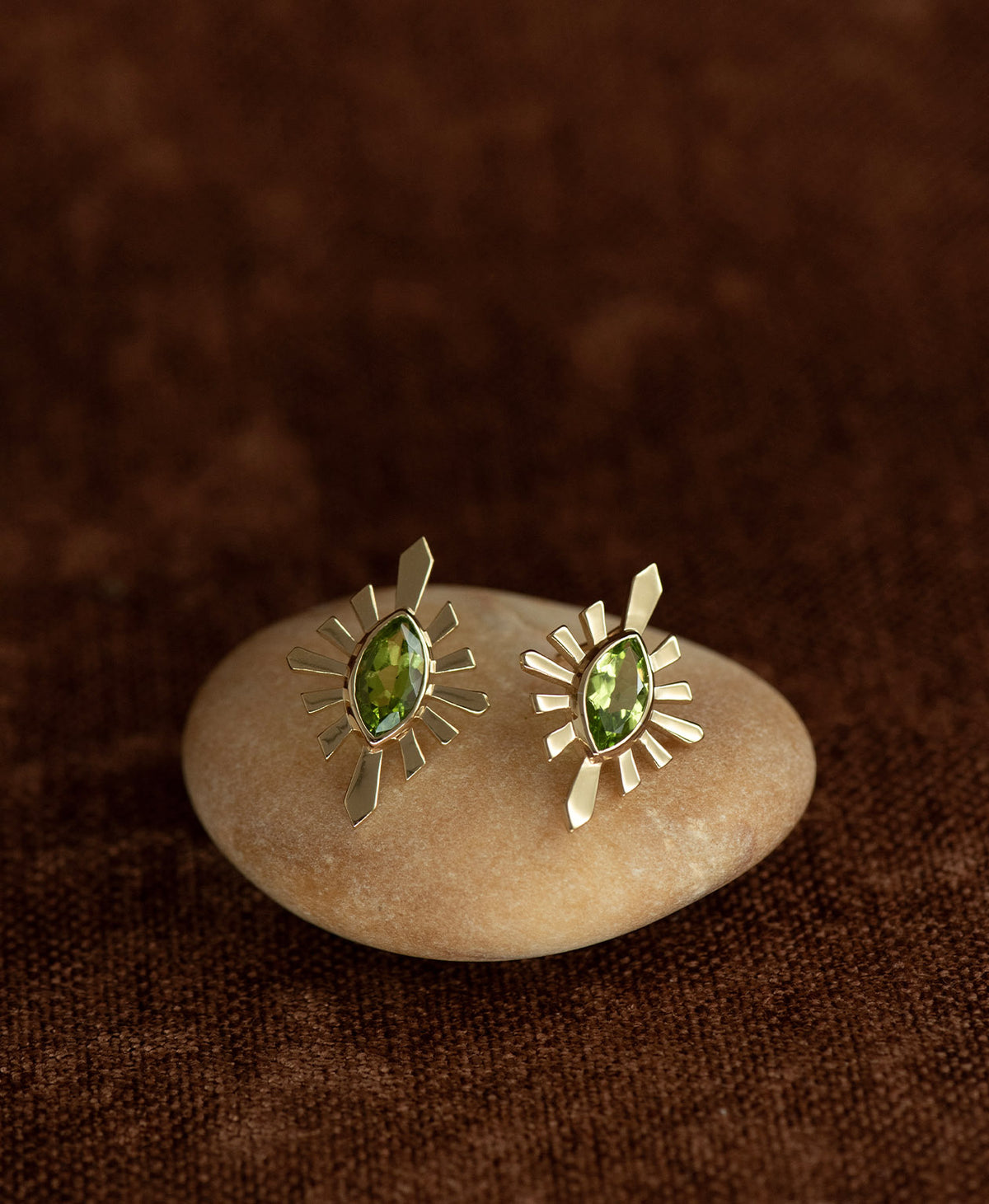 The Sister Studs, featuring a central marquise cut Peridot nestled within a silver sunburst frame, displayed on a smooth beige stone against a textured brown fabric background.