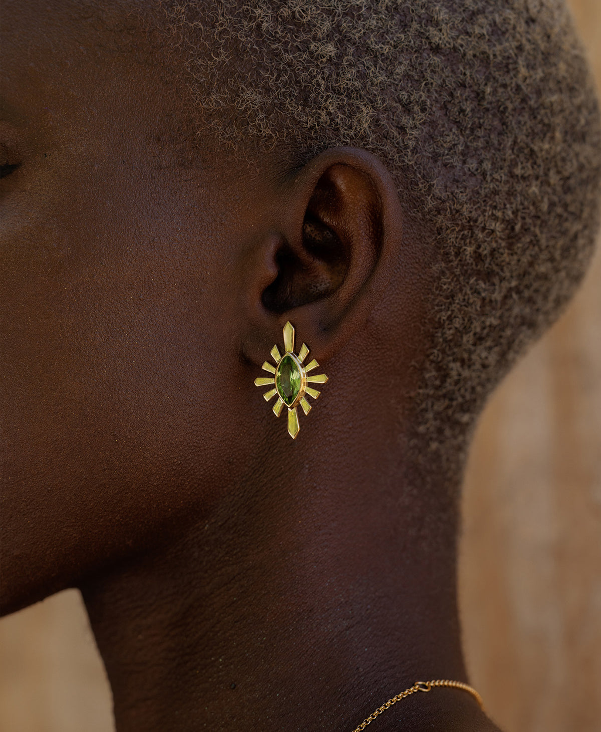 A close-up side profile of a person wearing the Sister Studs, featuring a marquise cut peridot in a gold linear sunburst frame. 