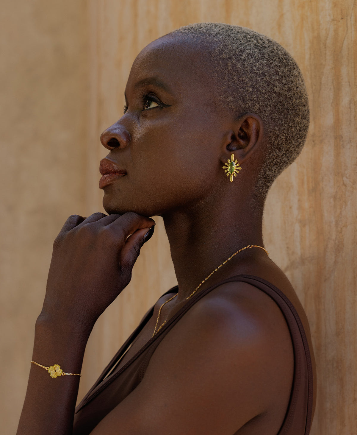 A side profile of a person wearing the Sister Studs, featuring a marquise cut peridot in a gold linear sunburst frame, paired with a gold bracelet. 