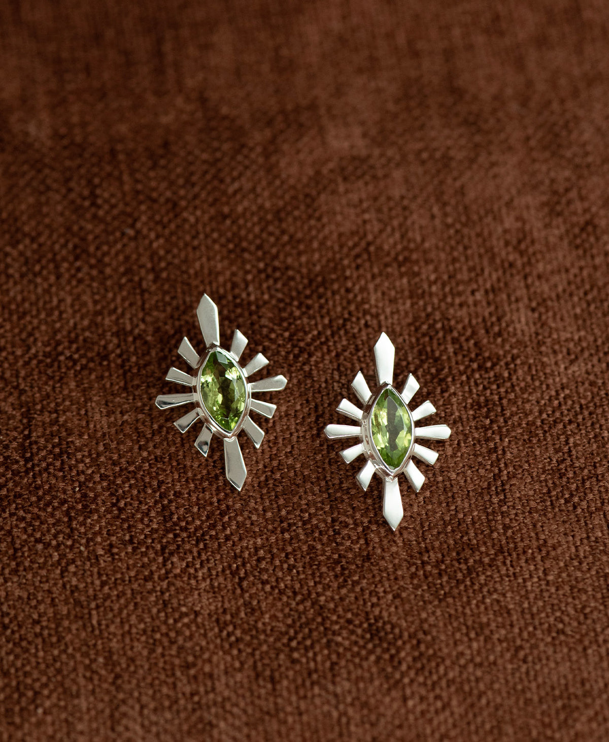 The Sister Studs, featuring a central marquise cut Peridot nestled within a silver sunburst frame, displayed against a textured brown fabric background.