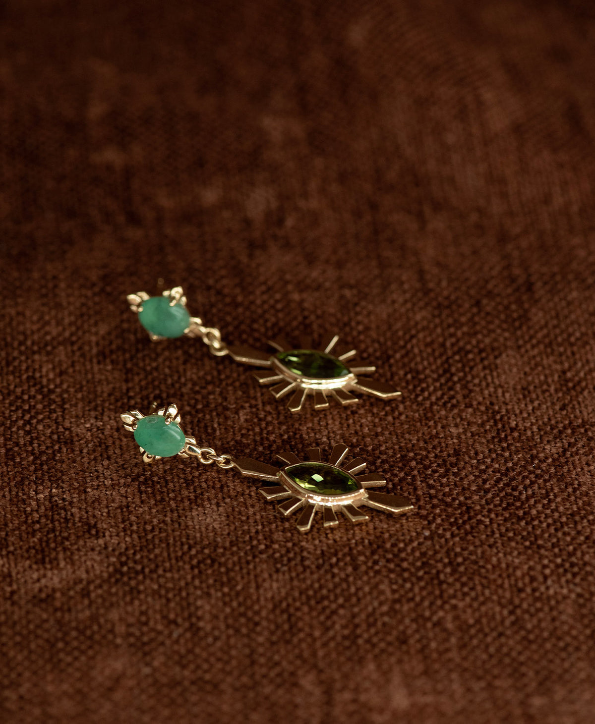 A pair of gold Sisterhood Earrings with a cabochon cut Emerald, set within a linear border, paired with a marquise cut Peridot nestled with a sunburst frame, displayed against a textured brown fabric background.