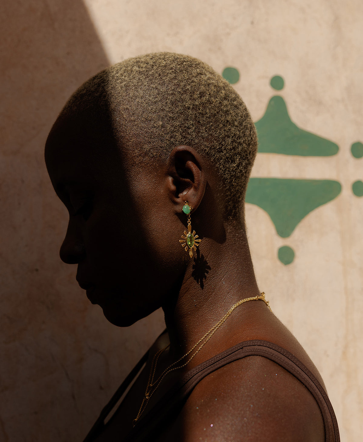 A close-up side profile of a person wearing the gold Sisterhood Earring, featuring a cabochon cut emerald set within a linear border, paired with a marquise cut peridot with a sunburst frame, and wearing 2 stacked gold necklace. 