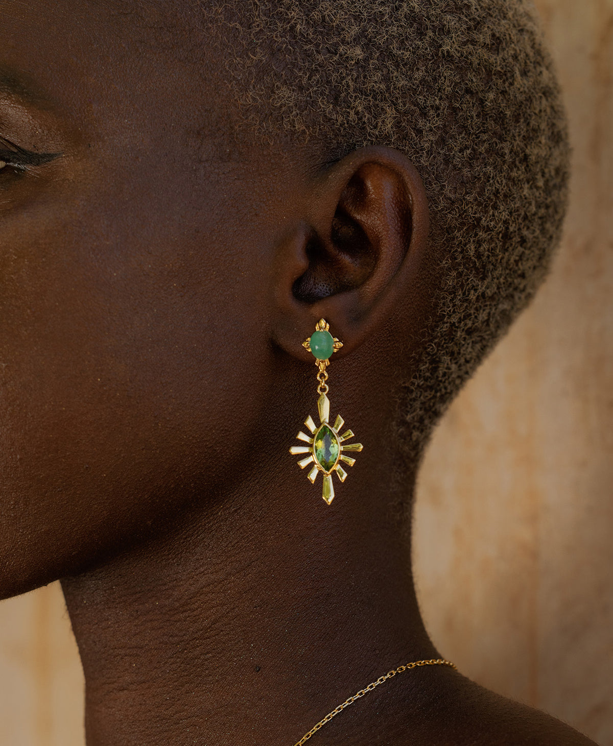 A close-up side profile of a person wearing the gold Sisterhood Earring, featuring a cabochon cut emerald set within a linear border, paired with a marquise cut peridot with a sunburst frame.