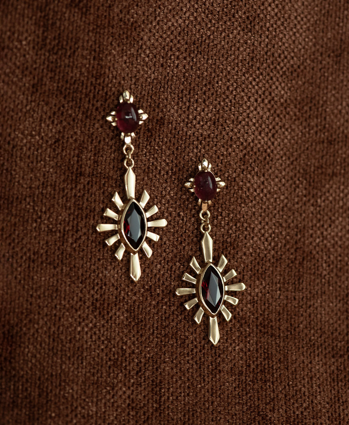 A pair of gold Sisterhood Earrings with a cabochon cut pink Tourmaline, set within a linear border, paired with a marquise cut Garnet nestled with a sunburst frame, hanging on a textured brown fabric background.