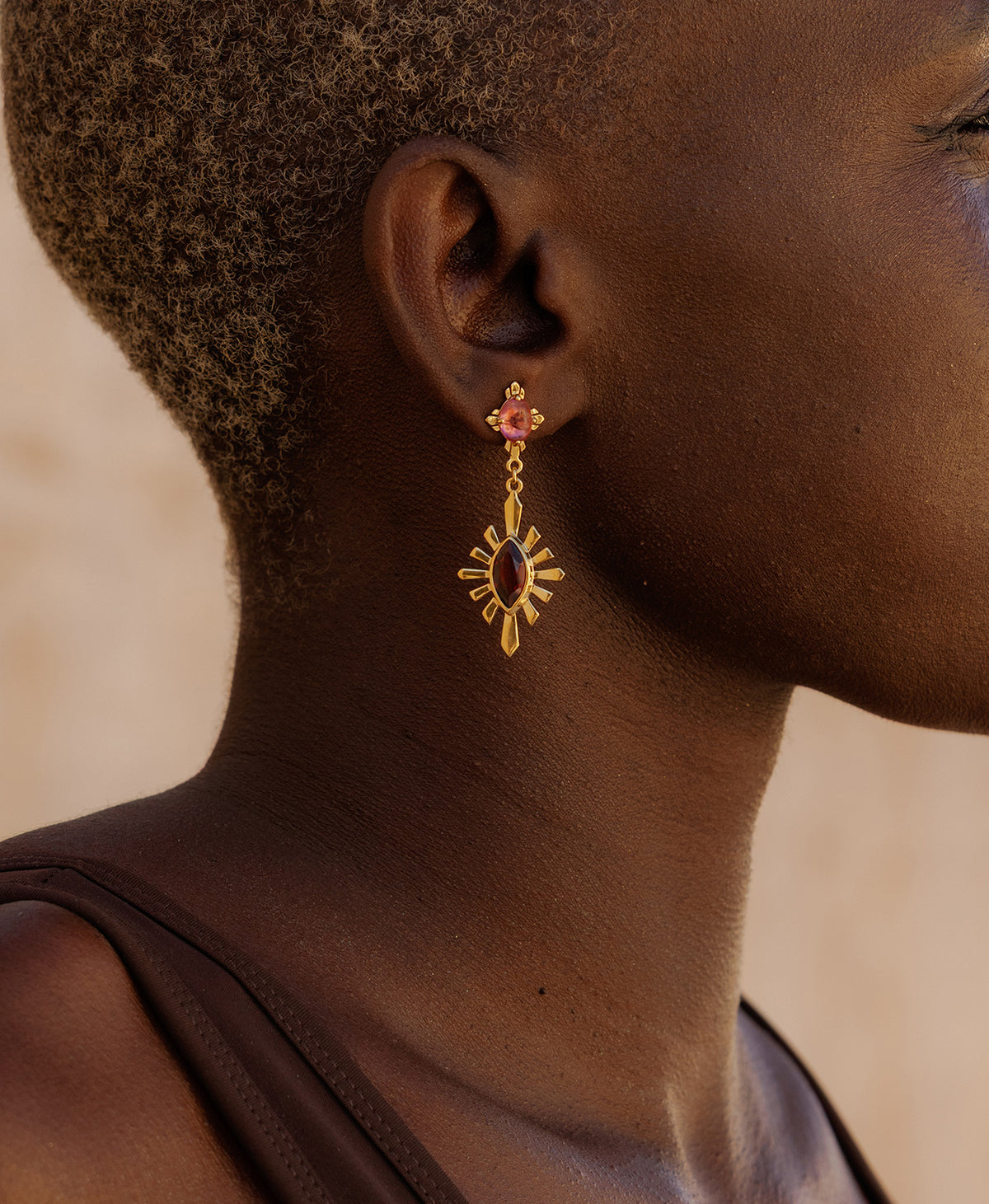 A close-up side profile of a person wearing the  Sisterhood Earring, featuring a cabochon cut pink Tourmaline is set within a linear border, paired with a marquise cut Garnet with a sunburst frame.