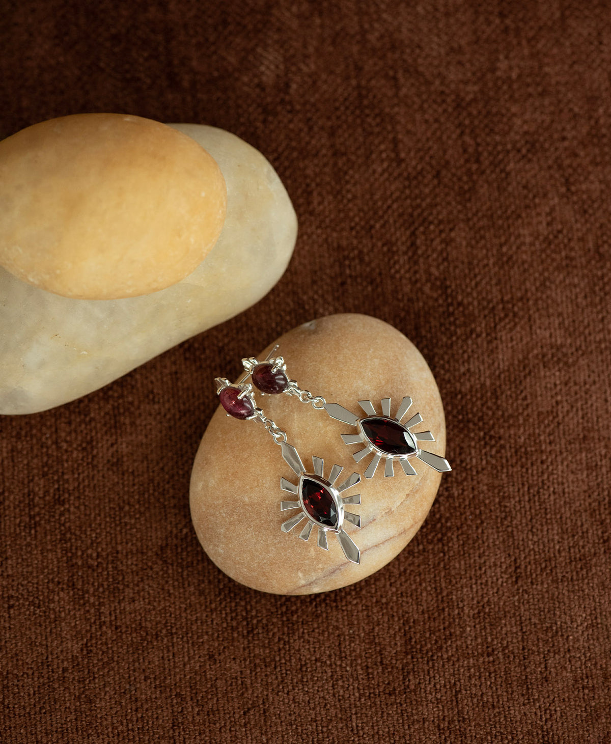 A pair of silver Sisterhood Earrings with a cabochon cut pink Tourmaline, set within a linear border, paired with a marquise cut Garnet nestled with a sunburst frame, displayed on a smooth beige stone against a textured brown fabric background.
