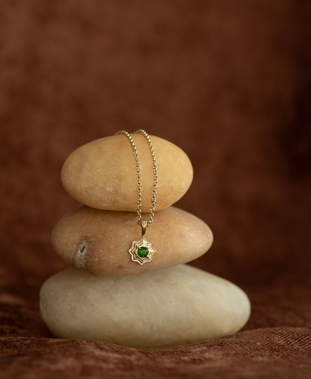 The Sitara Necklace featuring a gold star like pendant with concentric lines that radiate outwards from a Chrome Diopside resting at its centre, displayed over a stack of cream stones on a textured brown fabric background.