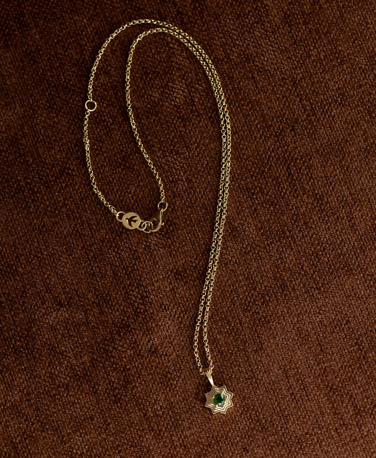 The Sitara Necklace featuring a gold star like pendant with concentric lines that radiate outwards from a Chrome Diopside resting at its centre, displayed on a textured brown fabric background.