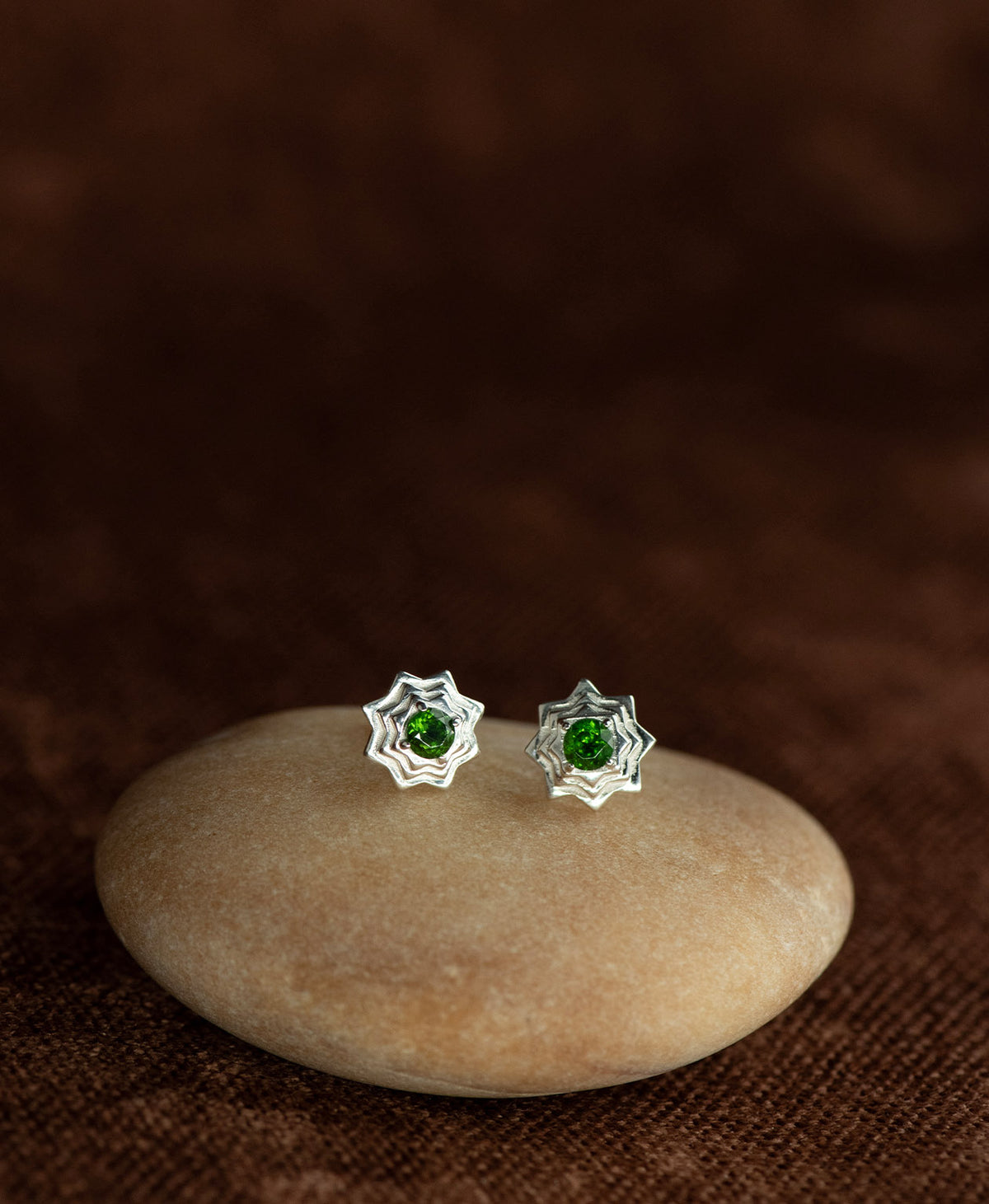 A pair of silver geometric star stud earrings with a central chrome diopside, displayed on a smooth cream coloured stone against a dark brown textured background.