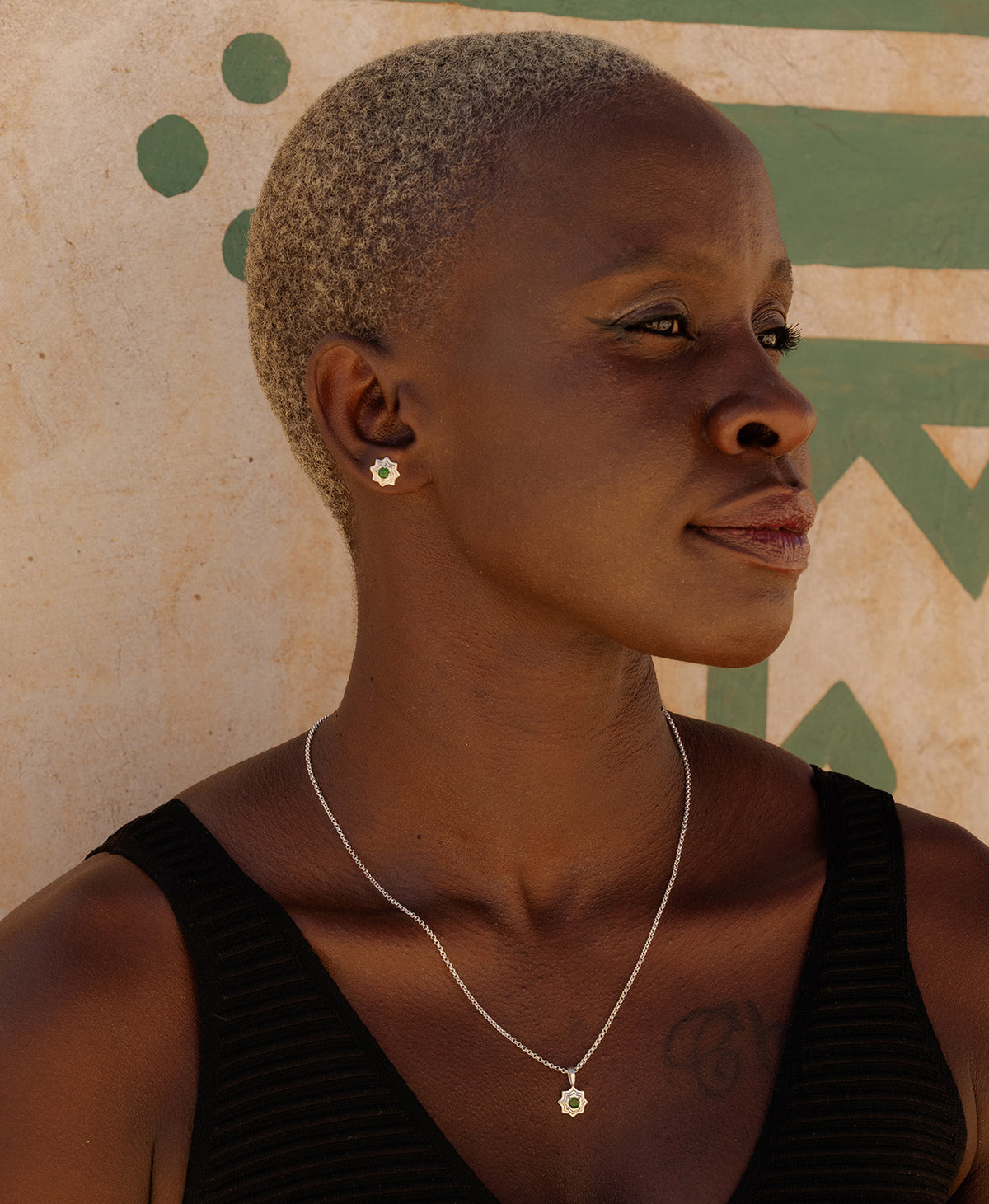 A profile of a person wearing the Sitara Studs, a silver star-like stud with a central green stone, paired with a matching silver pendant necklace. 