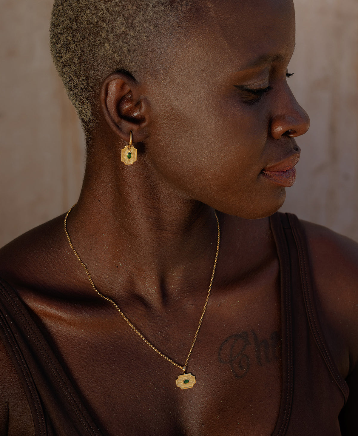 A person wearing the Soul Sister Necklace, a gold textured pendant with a central green stone, suspended from a fine belcher chain, paired with matching gold earrings. 
