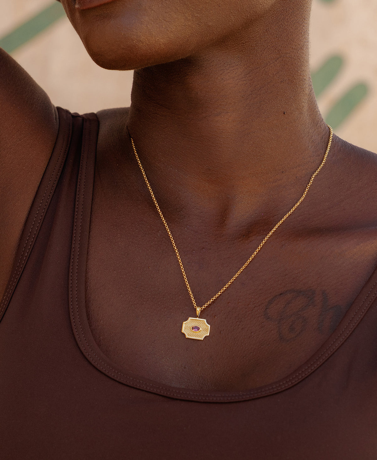 A close-up of a neck wearing the Soul Sister Necklace, a gold textured pendant with a central pink stone, suspended from a fine belcher chain.